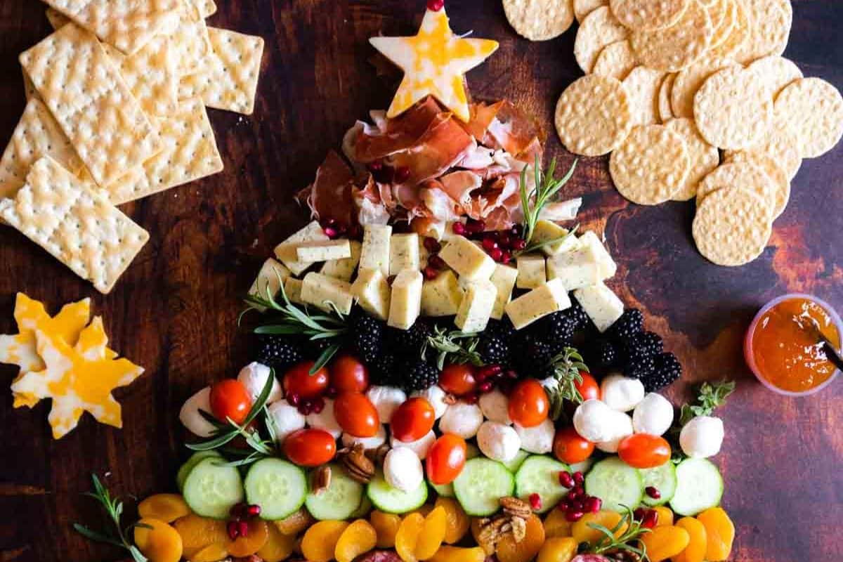 Charcuterie board arranged in a Christmas tree shape with meats, cheeses, fruits, and veggies-perfect for holiday appetizers-surrounded by crackers and a small bowl of jam on a wooden surface.