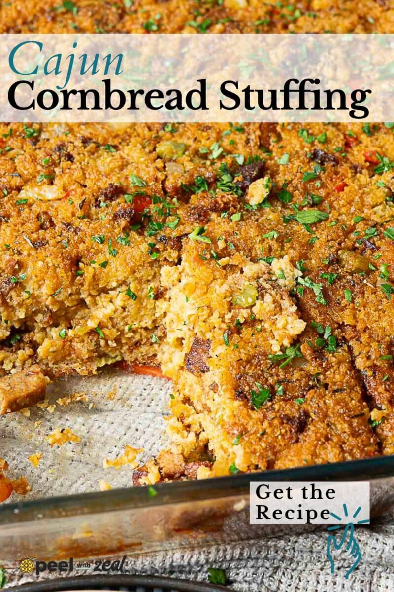 A rectangular dish filled with Cajun Cornbread Dressing, topped with herbs and a crispy golden crust, with a portion scooped out.