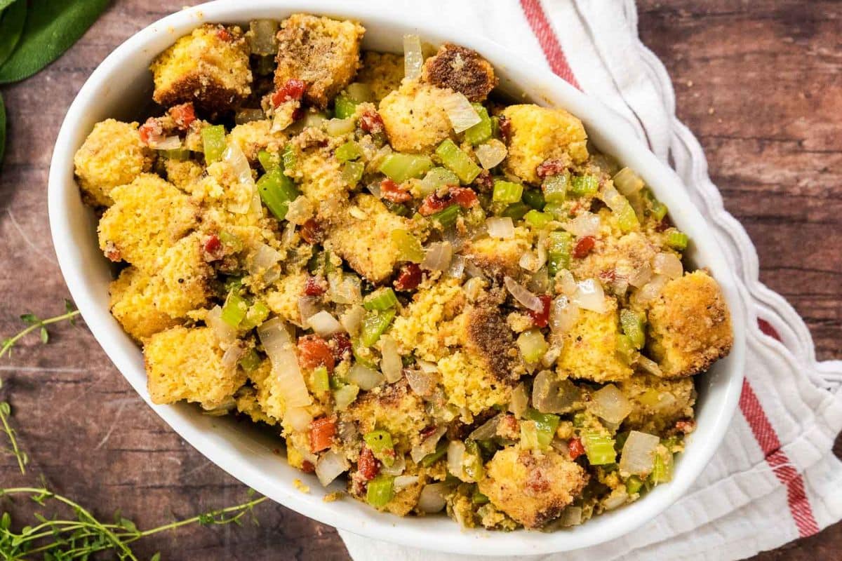 Oval dish filled with cornbread stuffing mixed with celery, onions, and bits of bacon, placed on a striped cloth on a wooden surface-one of the classic Thanksgiving sides perfect for your holiday table.