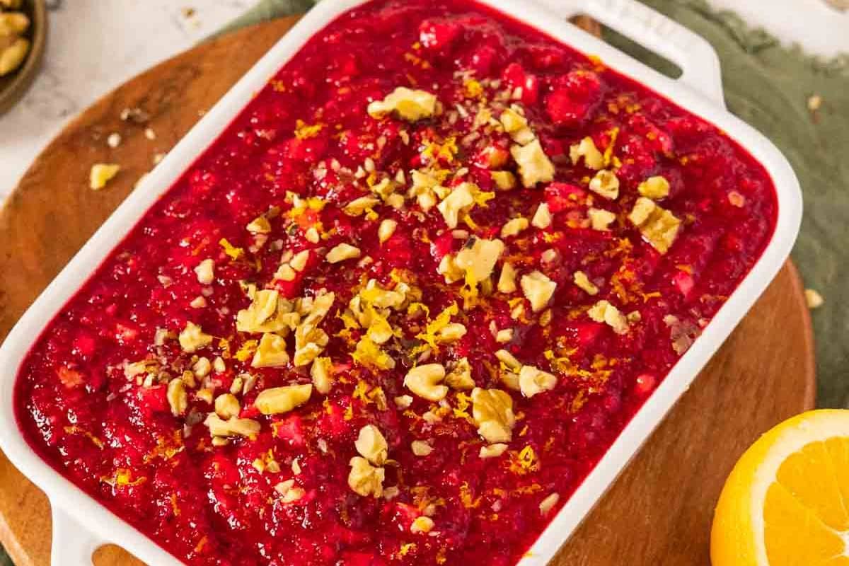 A rectangular white dish filled with cranberry salad, topped with chopped walnuts and citrus zest, sits on a wooden board next to a sliced orange-a festive addition to your holiday side dishes or Thanksgiving recipes.