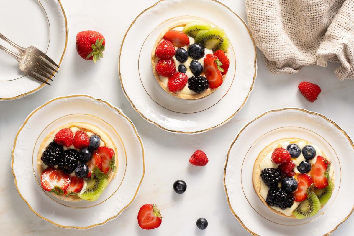Three plates each hold a fruit tart topped with strawberries, blackberries, blueberries, and kiwi slices. Loose berries and a beige cloth are also on the white surface.