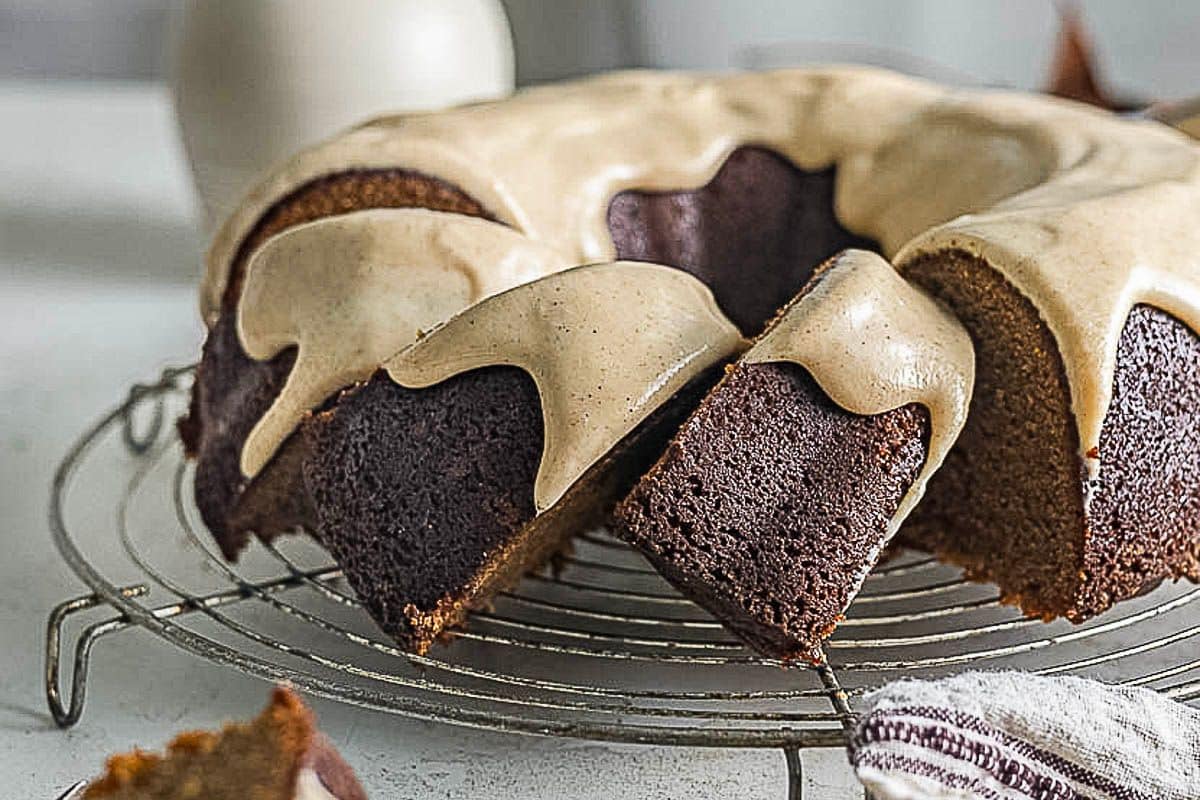 A bundt cake with beige icing is displayed on a wire rack, one slice partially removed-perfect for serving as gluten-free Thanksgiving desserts.