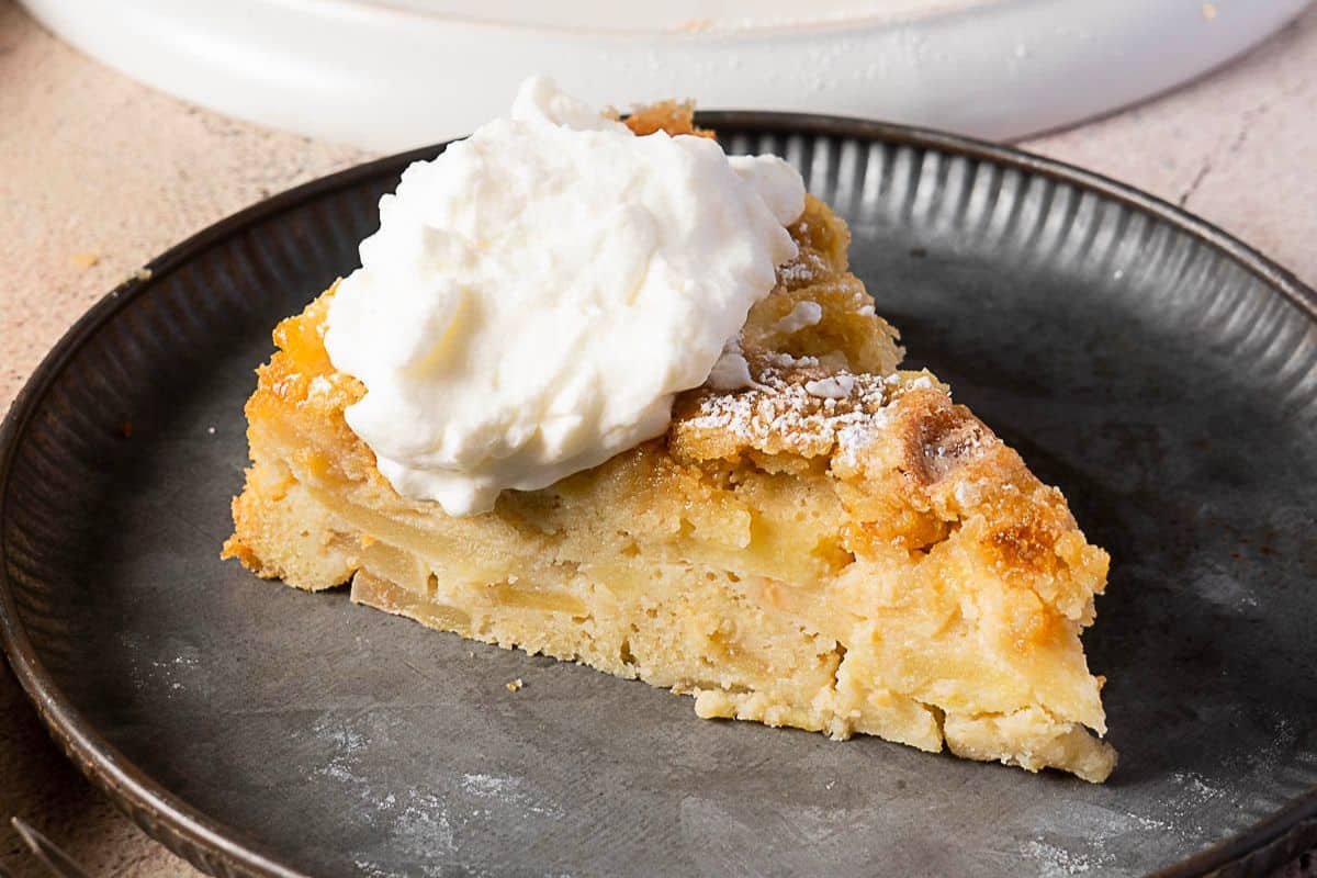 A slice of apple cake topped with a dollop of whipped cream on a dark plate-perfect for those seeking delicious gluten-free Thanksgiving desserts.