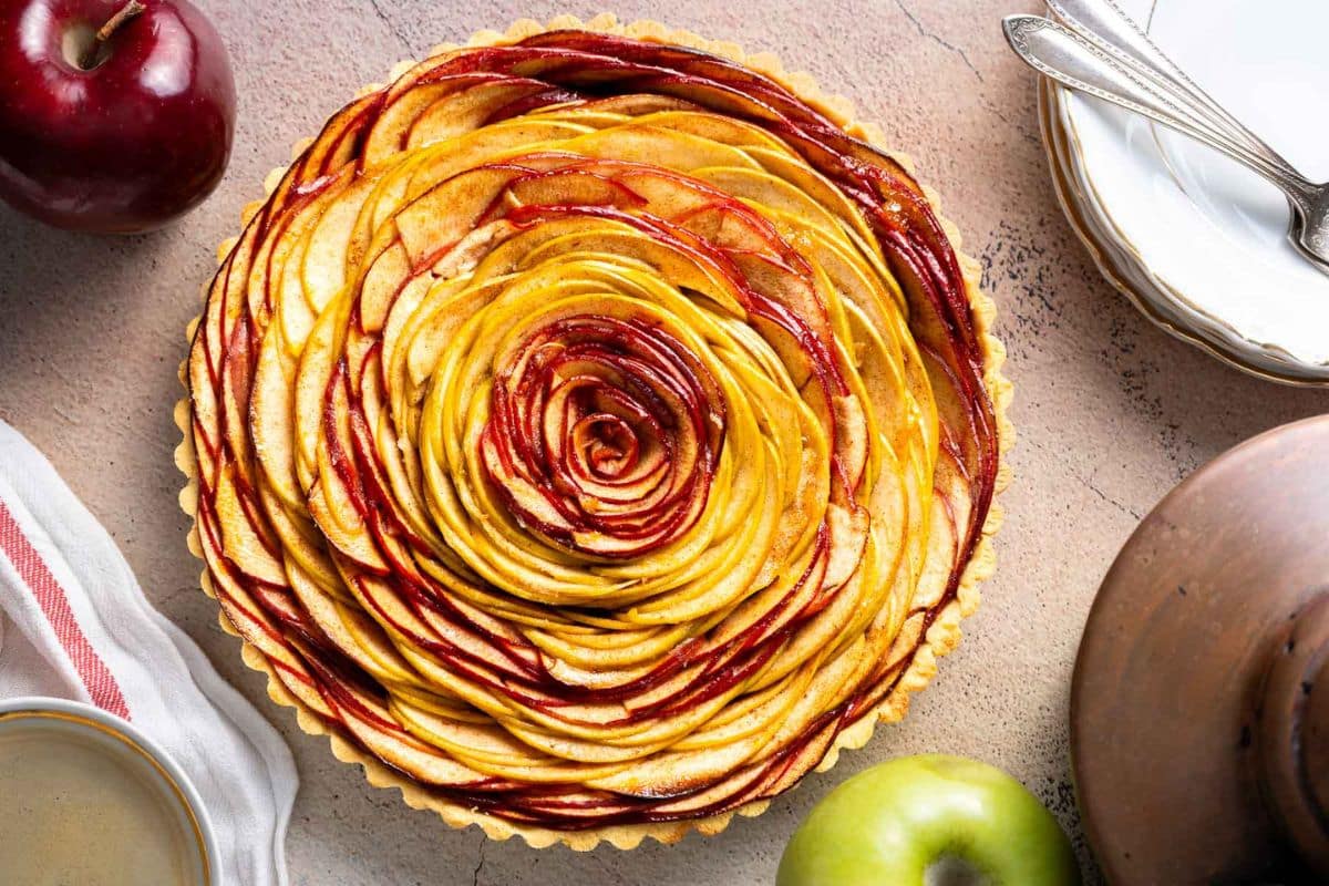 A baked apple tart with thinly sliced red and green apples arranged in concentric circles, surrounded by apples, plates, and cutlery on a light surface-an elegant choice for gluten-free Thanksgiving desserts.