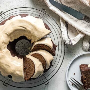 A bundt cake with light frosting sits on a wire rack; a few slices have been cut and served on small plates nearby with forks and a knife.