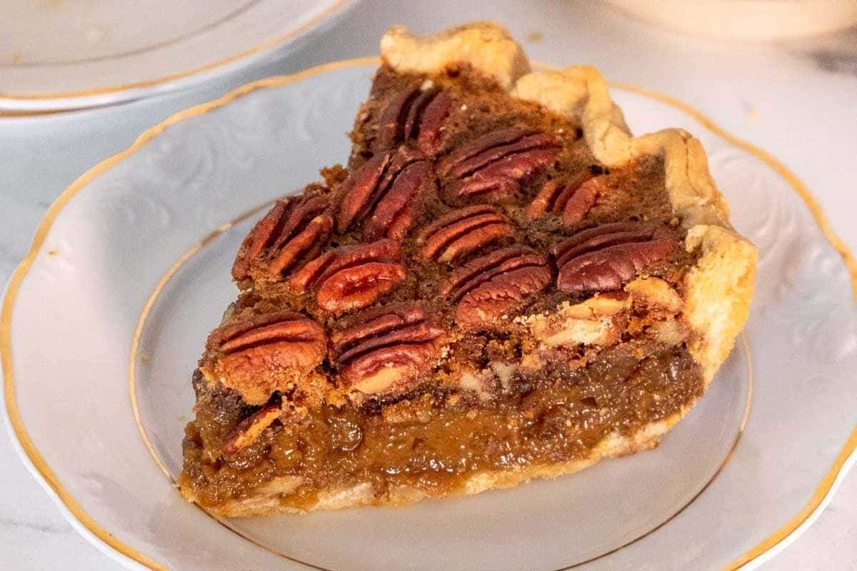 A slice of pecan pie sits on a white plate with a gold rim. This classic Thanksgiving Dessert features a flaky crust, rich brown filling, and whole pecans on top-perfect for your holiday spread.