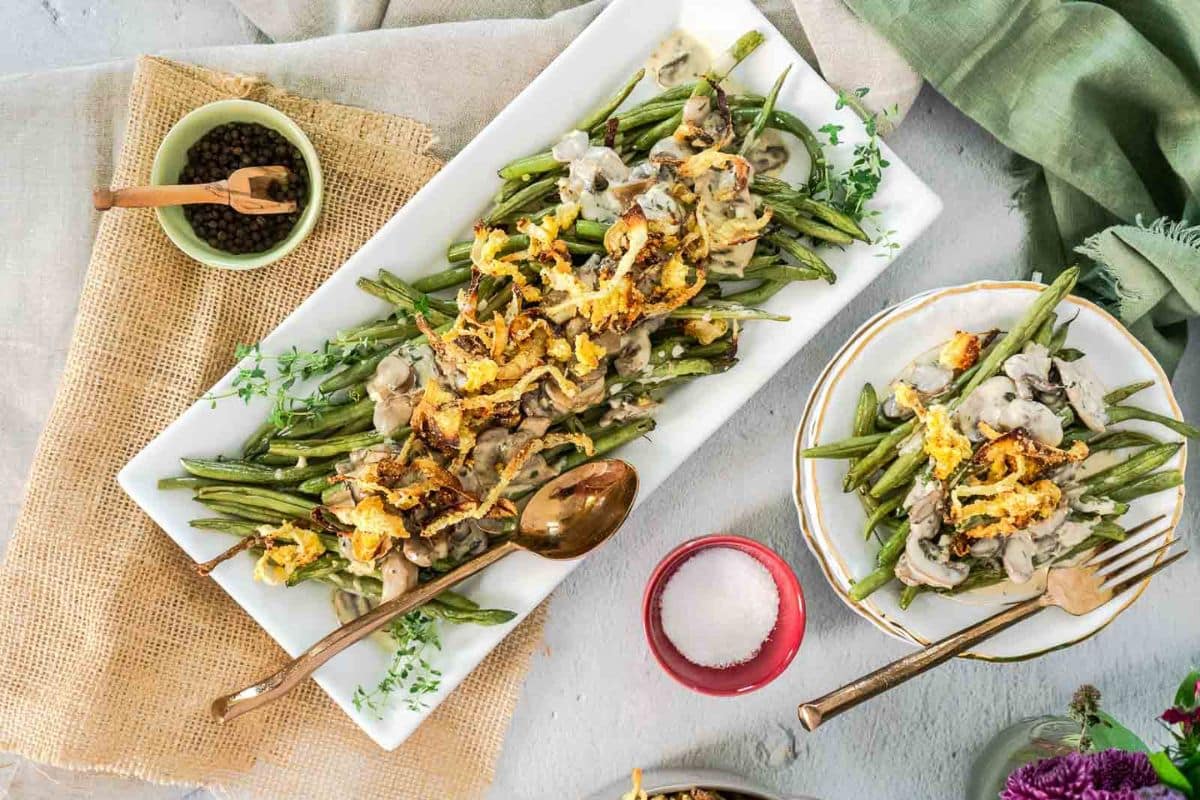 A rectangular white platter of green bean casserole, one of the classic Thanksgiving sides, topped with crispy onions and creamy sauce, with serving utensils beside a bowl of pepper and a dish of salt.
