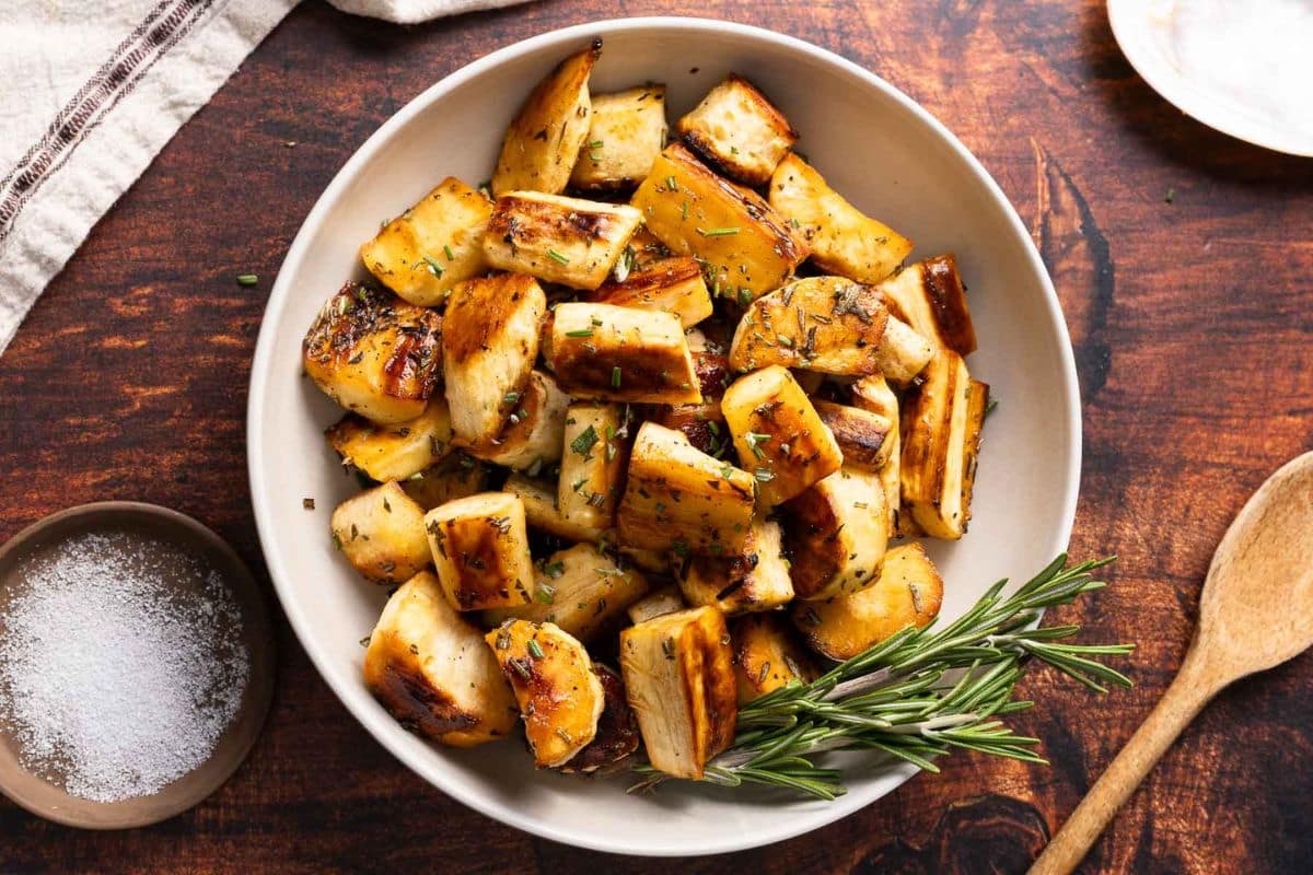 A bowl of roasted parsnips garnished with herbs sits on a wooden table next to a bowl of salt, a sprig of rosemary, and a wooden spoon-perfect for holiday side dishes or Thanksgiving recipes.
