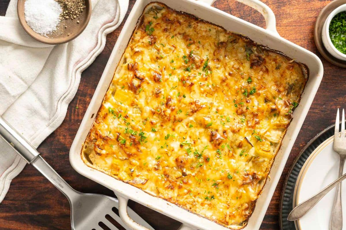 A baked casserole dish filled with cheesy scalloped potatoes, one of the best Thanksgiving sides, is garnished with chopped herbs and sits on a wooden table next to plates, utensils, and seasonings.