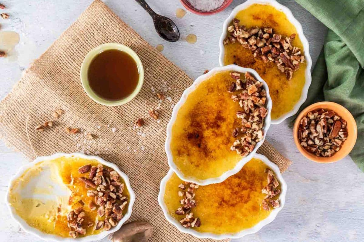 Four oval ramekins of cr&egrave;me br&ucirc;l&eacute;e topped with chopped pecans are arranged on a burlap cloth, alongside a small bowl of syrup and a bowl of pecans-perfect for gluten-free Thanksgiving desserts.