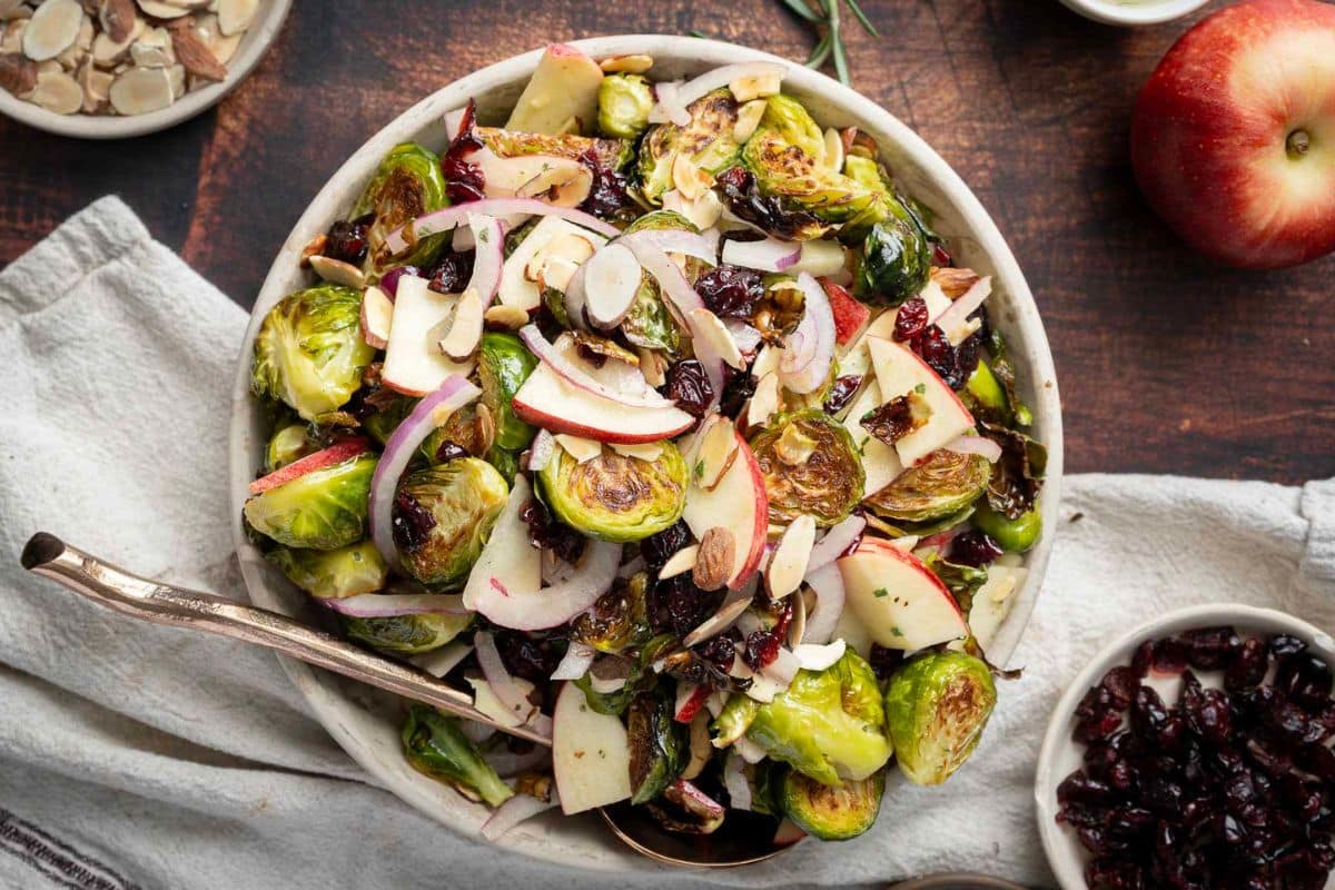 A bowl of salad with roasted Brussels sprouts, sliced apples, red onion, dried cranberries, and sliced almonds on a wooden table-perfect for holiday side dishes or Thanksgiving recipes.