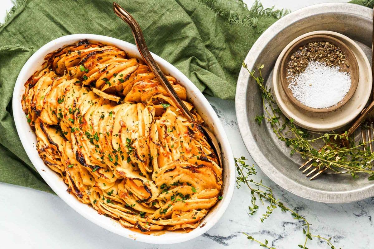 A baked dish of thinly sliced potatoes garnished with herbs sits next to a bowl containing salt, pepper, and fresh thyme on a marble surface-perfect as one of your Thanksgiving sides.