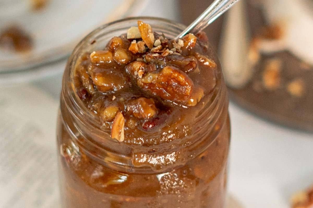 A glass jar filled with rich, brown pecan pie filling, topped with chopped pecans and a spoon inside-perfect for those seeking delicious Gluten-Free Thanksgiving Desserts.