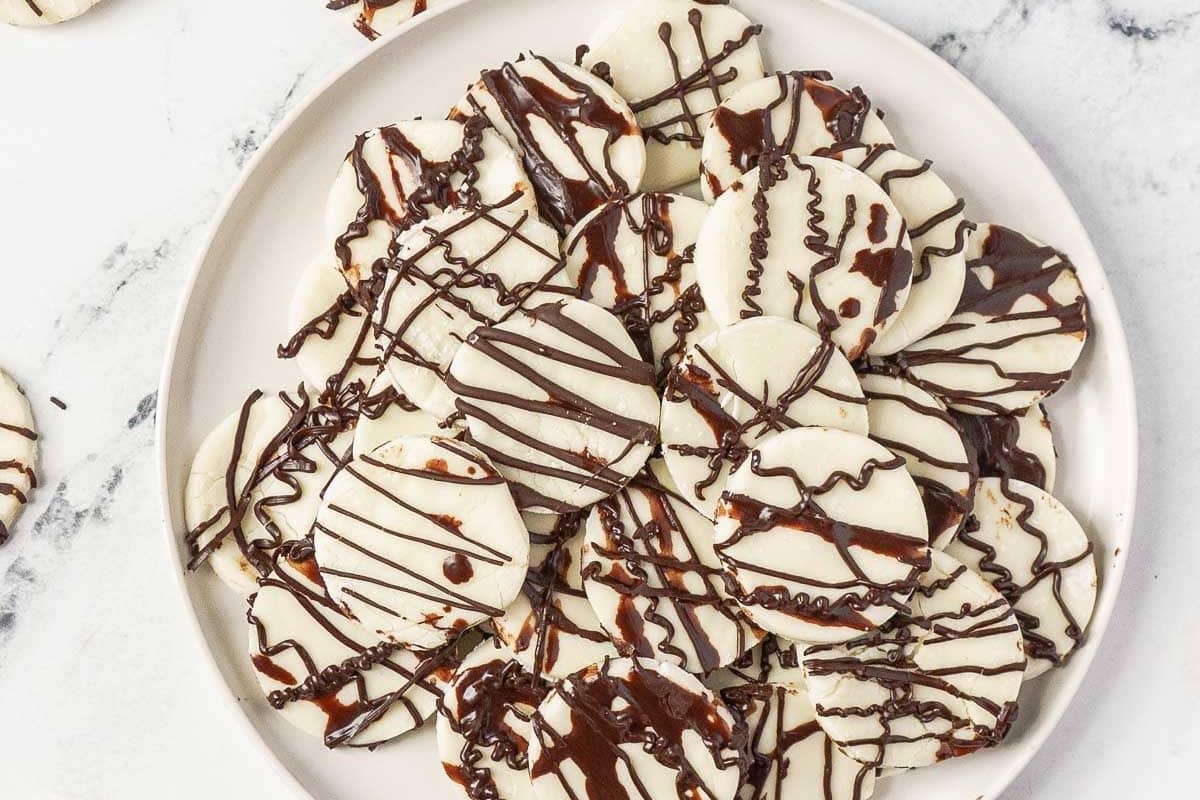 A plate of round cookies covered in white icing and drizzled with dark chocolate, arranged on a white plate.