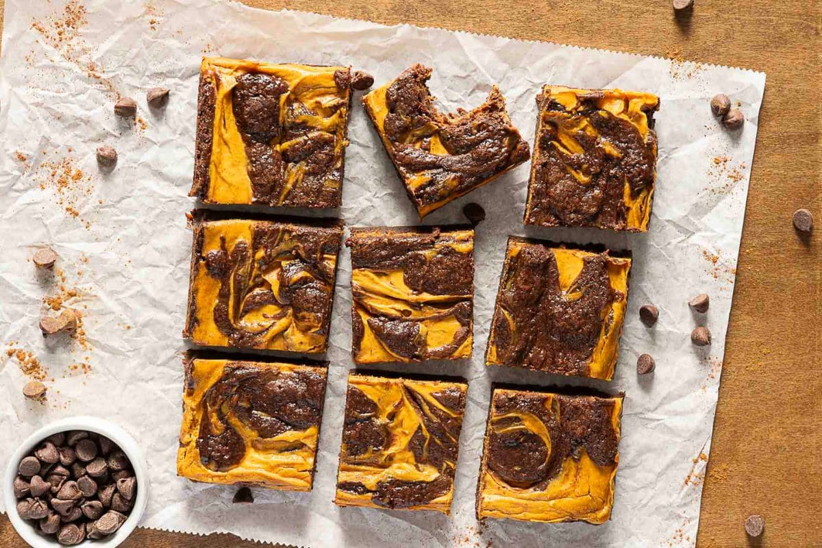 Nine pumpkin swirl brownies arranged on parchment paper, with one brownie slightly separated and partially eaten; chocolate chips are scattered beside a small bowl in the corner-perfect for gluten-free Thanksgiving desserts.
