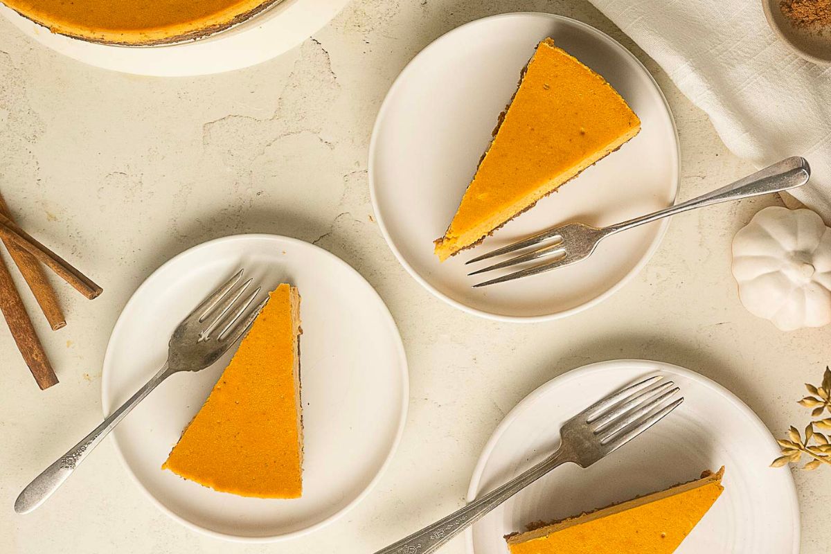 Three slices of pumpkin pie on white plates with forks, arranged on a light-colored surface beside a whole pie, cinnamon sticks, and a small decorative pumpkin-perfect for those seeking delicious gluten-free Christmas desserts.
