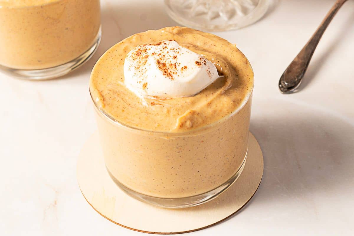 A glass cup filled with creamy pumpkin mousse, a perfect choice for gluten-free Thanksgiving desserts, topped with whipped cream and a sprinkle of spice, sits on a white coaster.