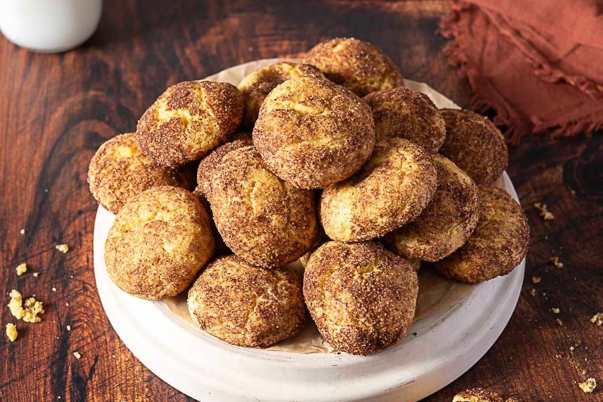 A plate of round, golden-brown cookies coated in cinnamon sugar sits on a wooden surface with a brown napkin nearby.