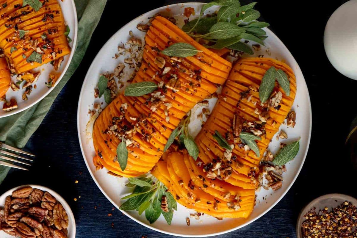 Two halves of hasselback butternut squash garnished with sage and chopped pecans are served on a white plate-a delicious highlight among Thanksgiving sides.