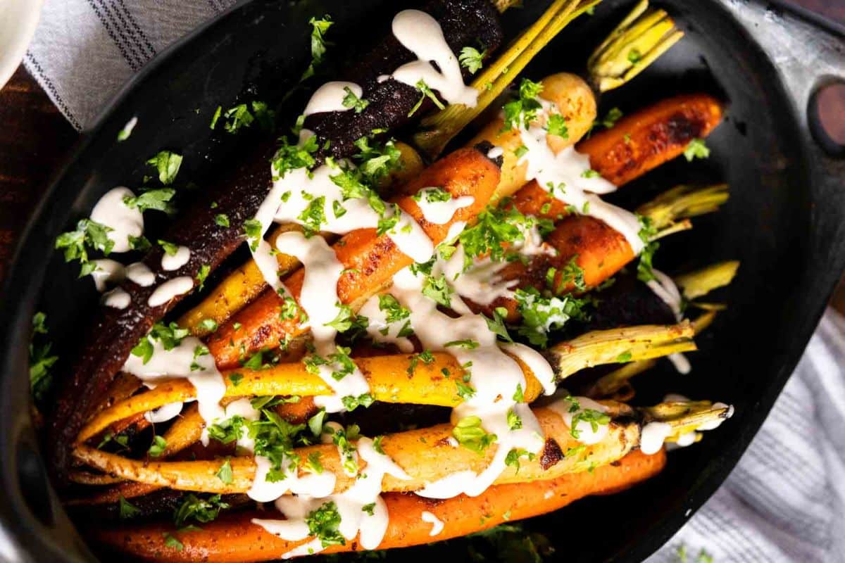 Roasted multicolored carrots topped with a creamy white sauce and chopped fresh herbs, served in a black dish-one of the perfect Thanksgiving sides for your holiday table.