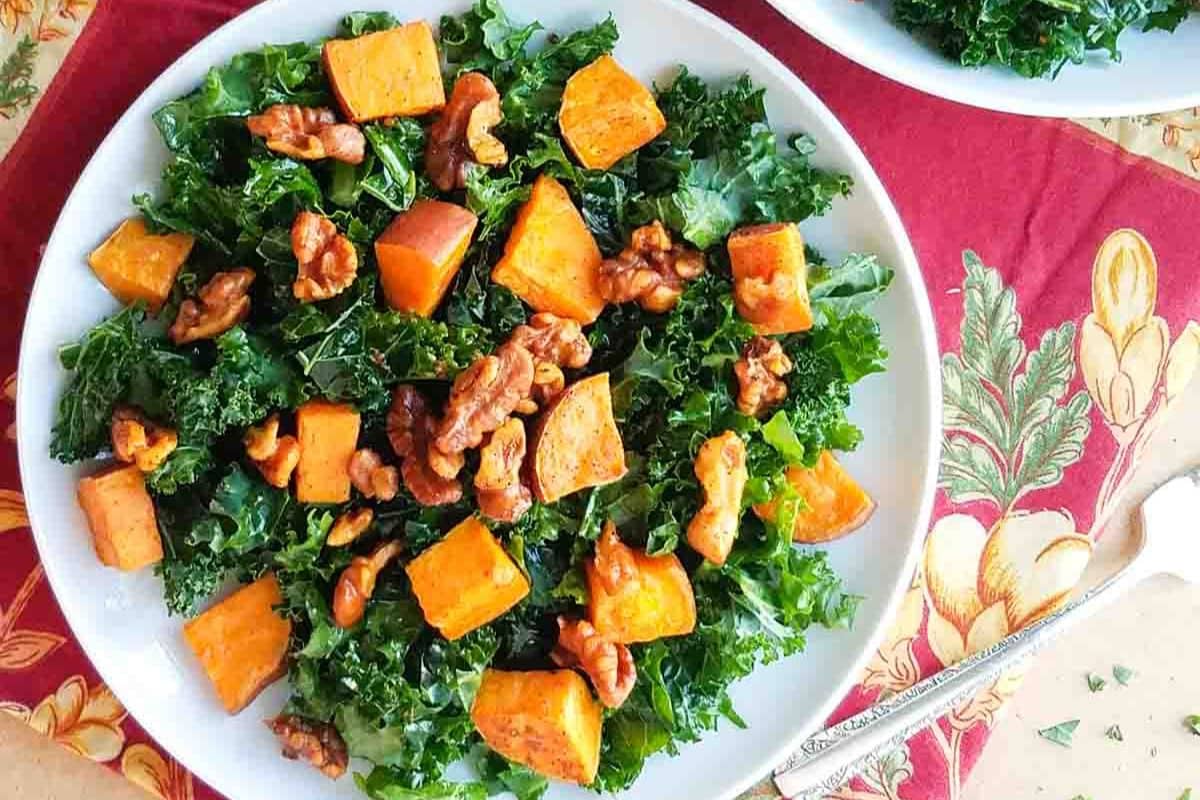A plate of kale salad topped with roasted sweet potato cubes and walnuts, placed on a red decorative tablecloth next to a fork-perfect for Thanksgiving sides or festive side dishes.