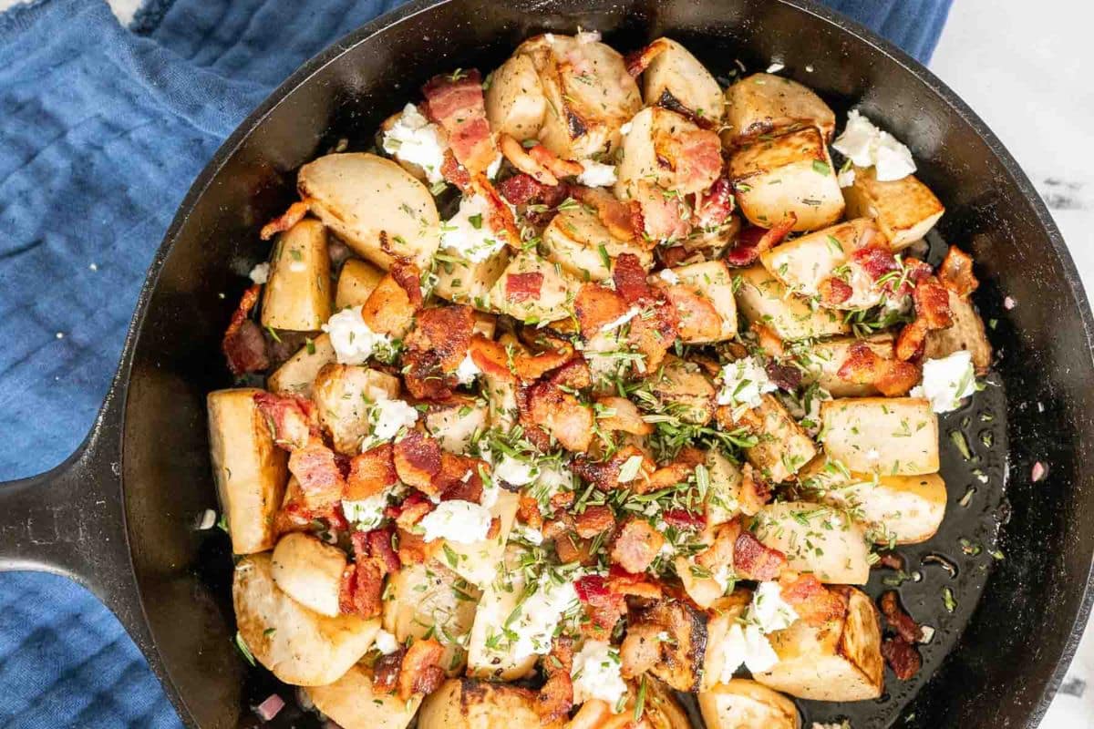 A skillet filled with roasted potatoes, crumbled bacon, goat cheese, and herbs rests on a blue cloth-delicious inspiration for Thanksgiving sides or festive Thanksgiving recipes.