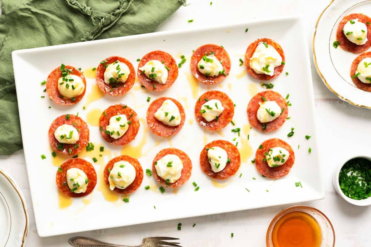 Rectangular white plate with round slices of pepperoni topped with dollops of cheese or cream, sprinkled with chopped chives, and drizzled with oil-perfect for easy last minute appetizers at any holiday gathering.