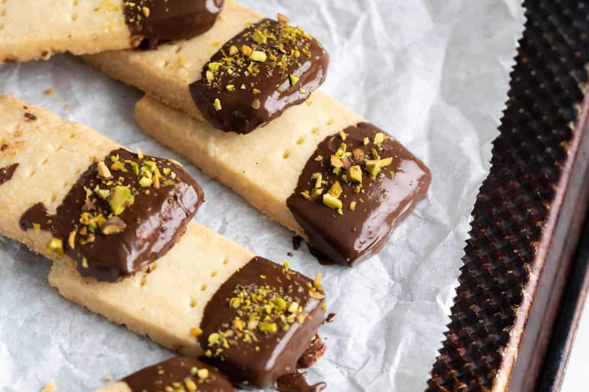 Rectangular shortbread cookies dipped in chocolate and sprinkled with chopped pistachios, arranged on parchment paper.