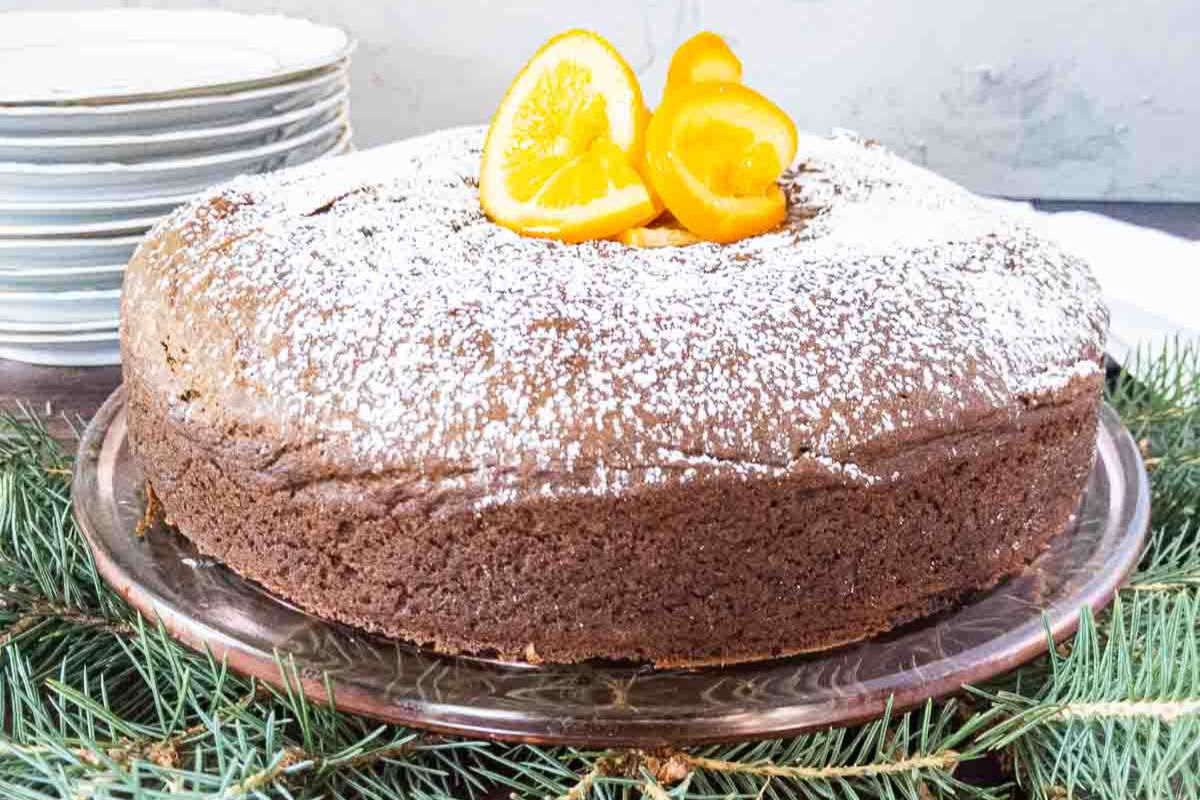 A round chocolate cake, perfect for Christmas Desserts, is topped with powdered sugar and orange slices, displayed on a glass plate with evergreen branches. Plates are stacked in the background.