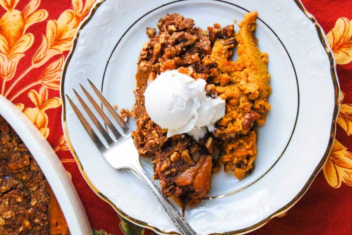 A serving of pumpkin crisp, a delightful addition to Thanksgiving sides, is topped with vanilla ice cream on a decorative white plate with a fork, set on a red patterned tablecloth.