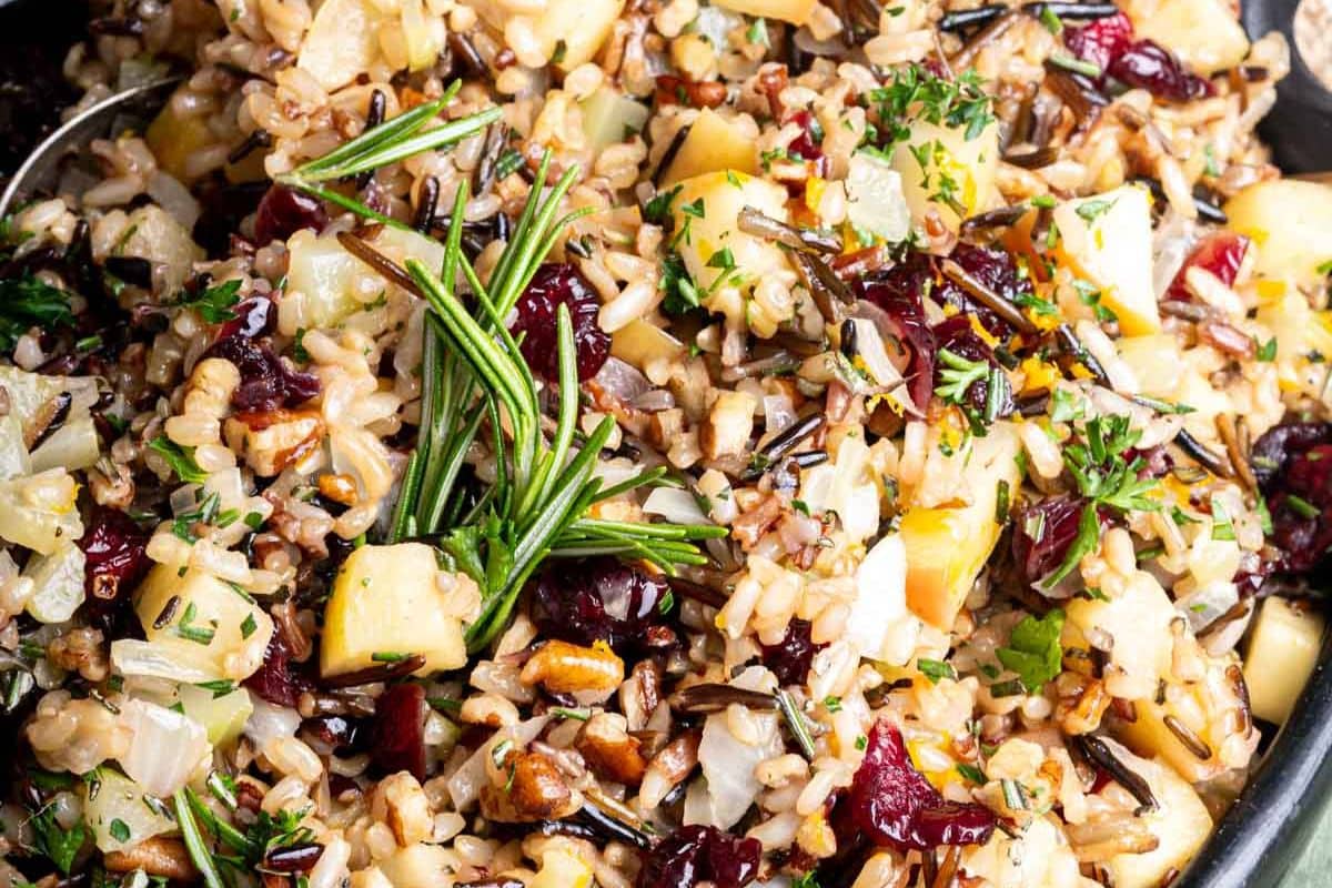 A close-up of wild rice pilaf mixed with diced apples, cranberries, pecans, chopped herbs, and a fresh rosemary sprig-perfect for Thanksgiving recipes or as one of your favorite holiday side dishes.