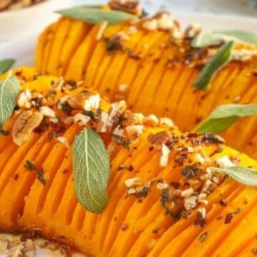 Sliced roasted butternut squash topped with chopped pecans, fresh sage, and herbs—an inviting addition to the Holiday Table and perfect for Gluten-Free Thanksgiving Sides. Arranged beautifully on a white plate.