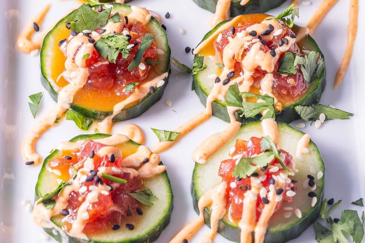 Slices of cucumber topped with diced raw fish, chopped herbs, sesame seeds, and a creamy orange sauce make elegant holiday appetizers, beautifully arranged on a white plate for your next party.