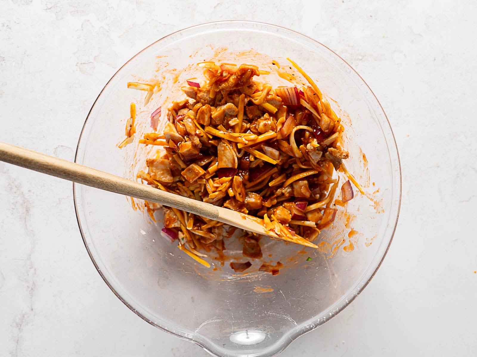 A glass mixing bowl contains shredded cheese, chopped red onions, and sauce being mixed with a wooden spoon.