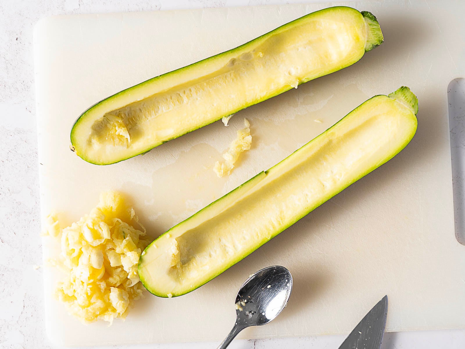 A zucchini cut in half lengthwise with the inside scooped out, placed on a cutting board next to a spoon, knife, and zucchini flesh.