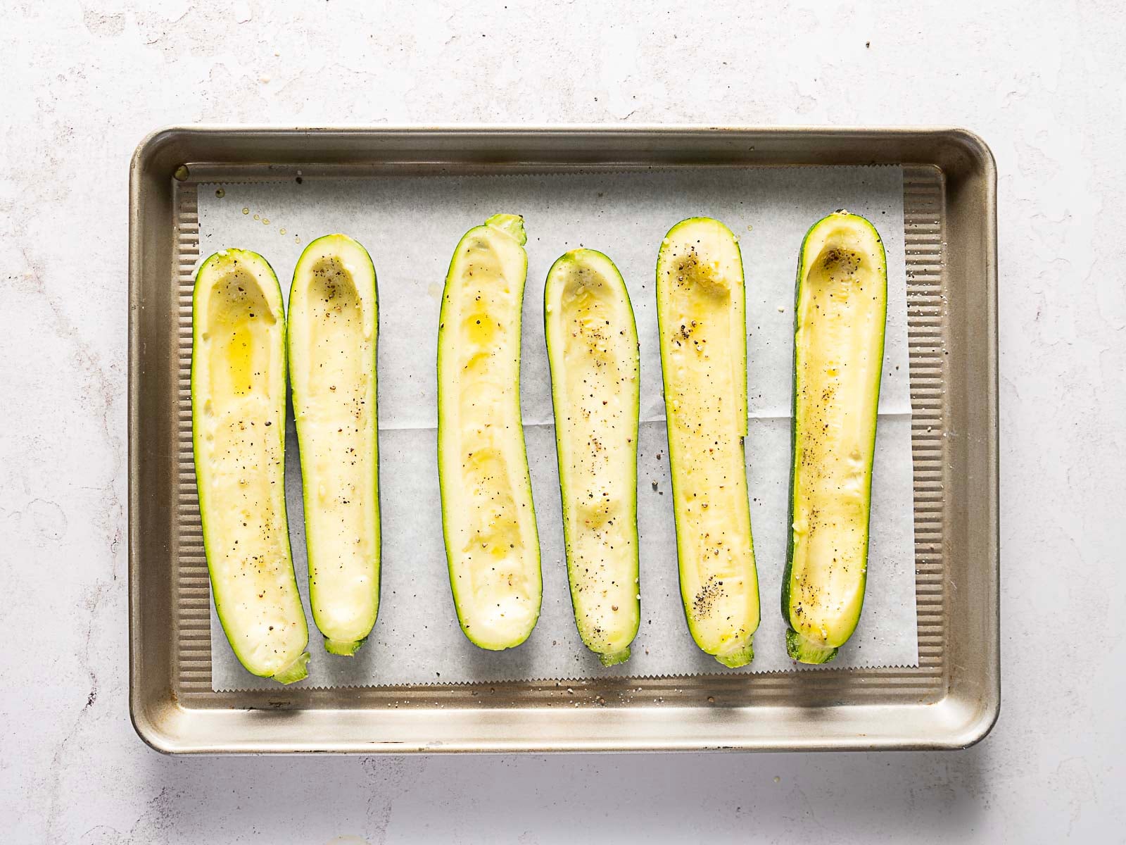 Six halved and hollowed zucchinis arranged in a row on a parchment-lined baking sheet, seasoned with oil, salt, and pepper.