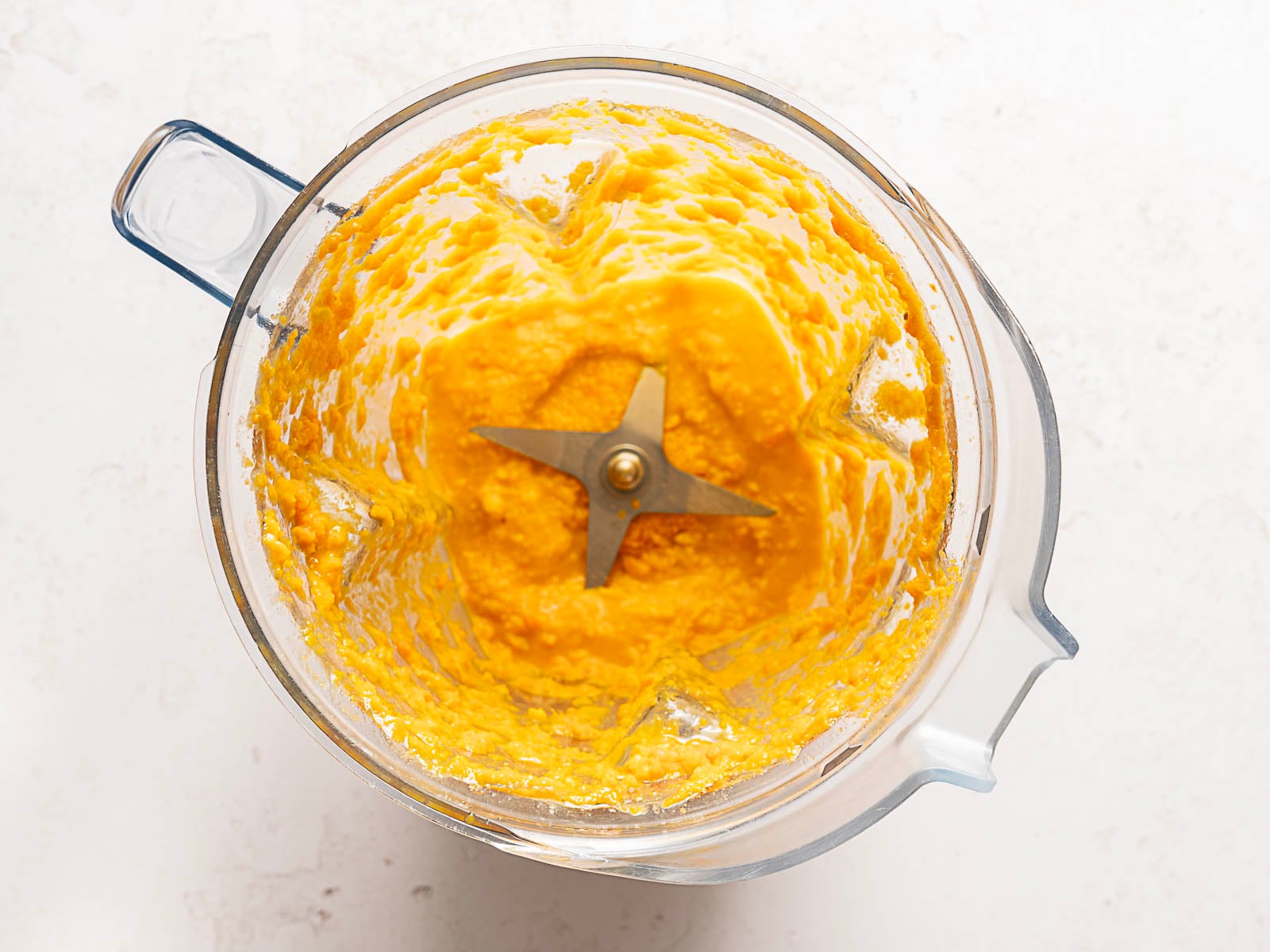 A top-down view of a blender with a stainless steel blade, containing a thick, smooth, bright orange mixture against a white background.