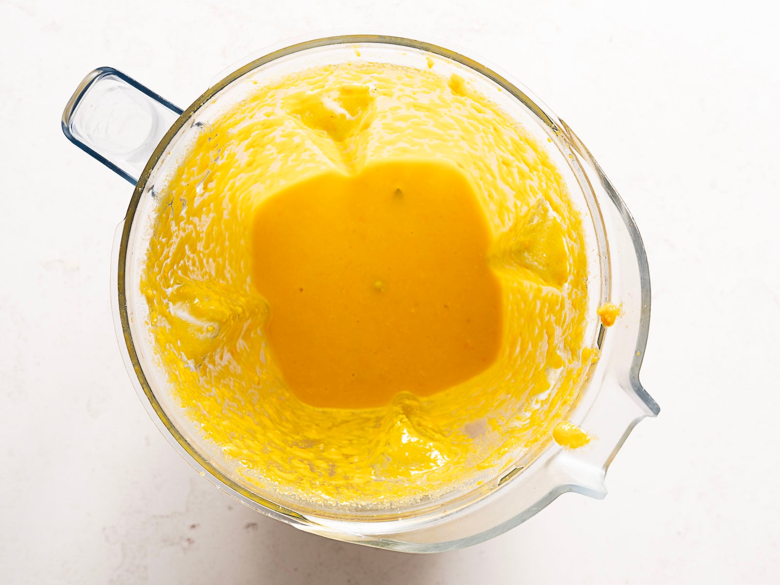 A top-down view of a glass blender pitcher containing a smooth, bright yellow mixture. Some of the mixture is splattered on the sides of the pitcher, and the blender is placed on a white surface.