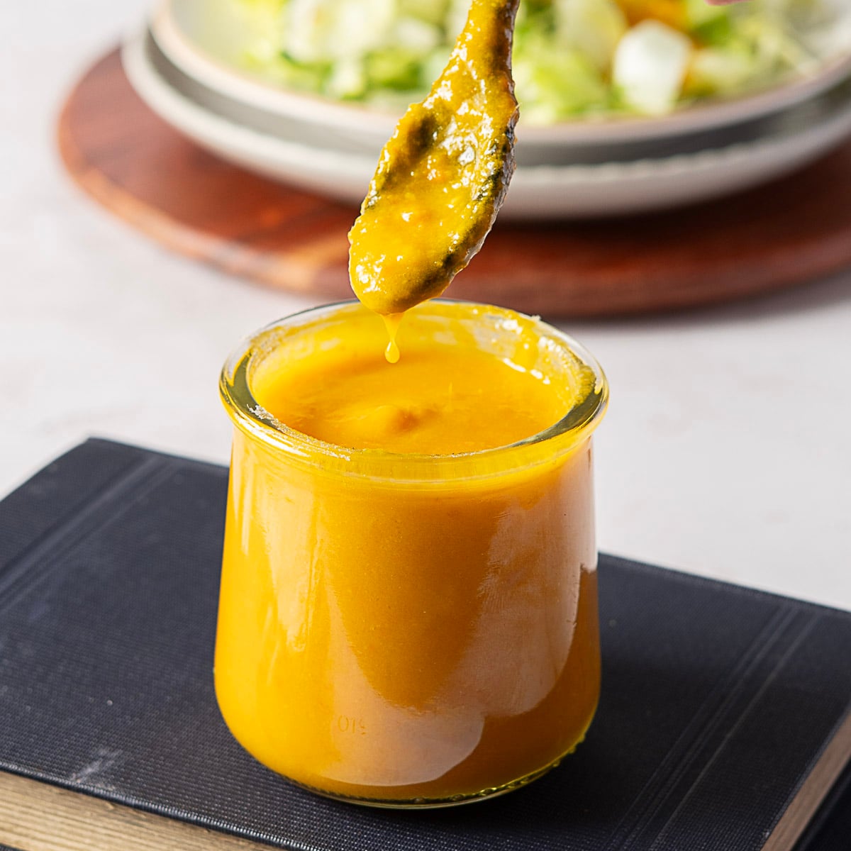 A glass jar filled with thick, yellow-orange dressing sits on a closed book. A spoon above the jar drips some of the dressing back in, and a plate of salad is blurred in the background.