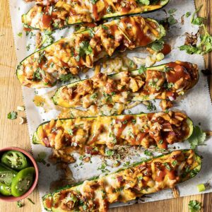 Baked zucchini halves stuffed with cheese, sauce, and herbs on parchment paper, with sliced jalapenos on the side.