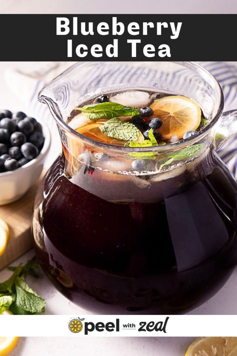 A glass pitcher filled with blueberry iced tea garnished with lemon slices, blueberries, mint leaves, and ice cubes. A bowl of fresh blueberries and some mint leaves are visible nearby. The text reads “Blueberry Iced Tea.”.