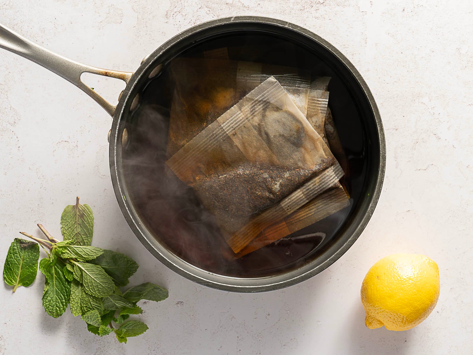 A saucepan filled with hot water and several tea bags, with steam rising. Fresh mint leaves and a whole lemon are placed on the countertop next to the saucepan.