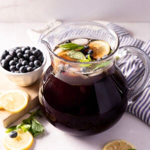 A glass pitcher filled with dark purple blueberry iced tea, garnished with lemon slices, blueberries, and mint. A bowl of blueberries, sliced lemons, and mint leaves are nearby, next to a blue-striped cloth.