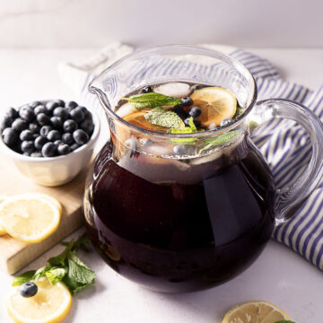 A glass pitcher filled with dark purple blueberry iced tea, garnished with lemon slices, blueberries, and mint. A bowl of blueberries, sliced lemons, and mint leaves are nearby, next to a blue-striped cloth.