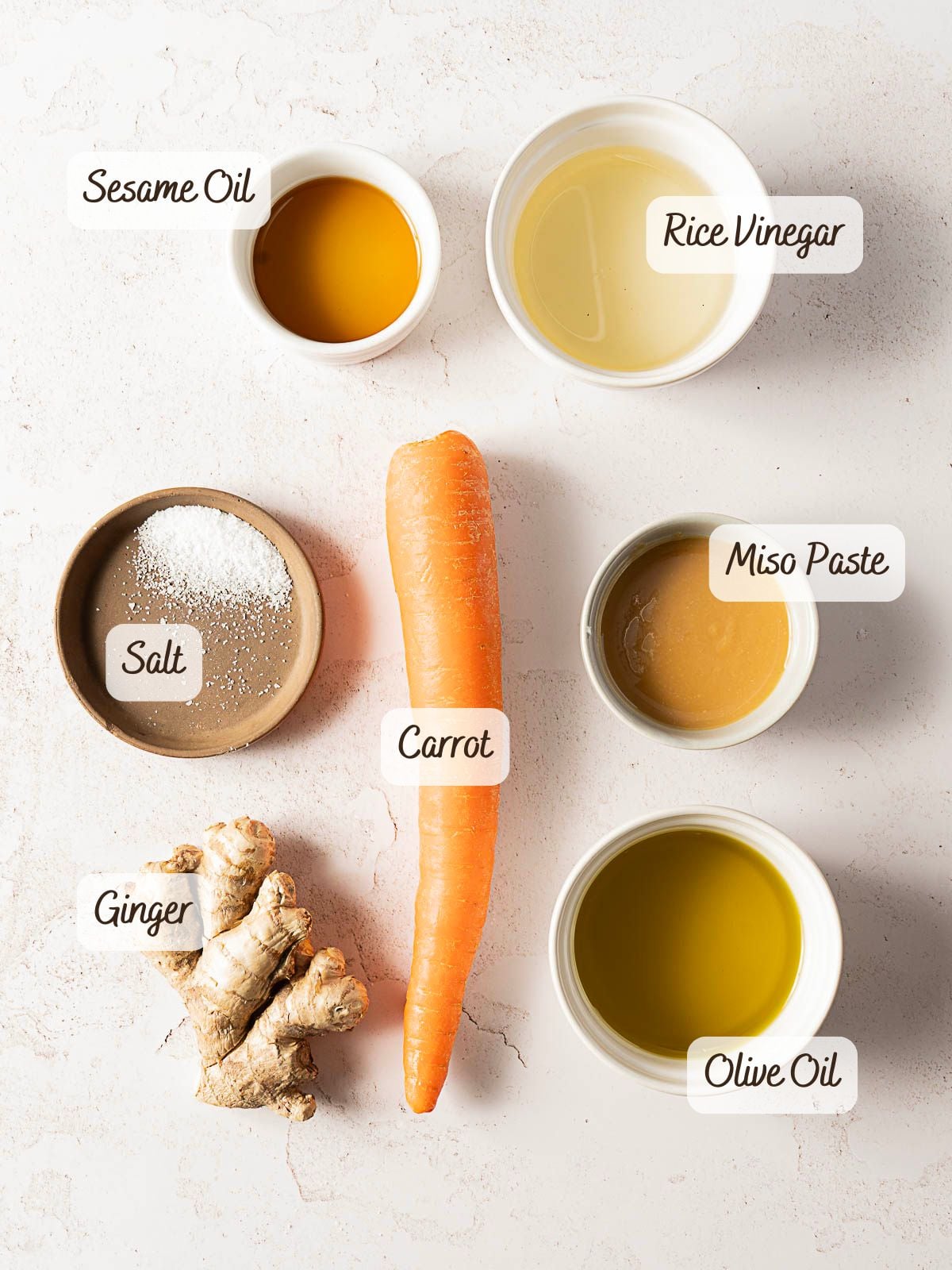 A flat lay of six labeled ingredients on a white surface: a carrot, a piece of ginger, a bowl of salt, and small bowls containing sesame oil, rice vinegar, miso paste, and olive oil.