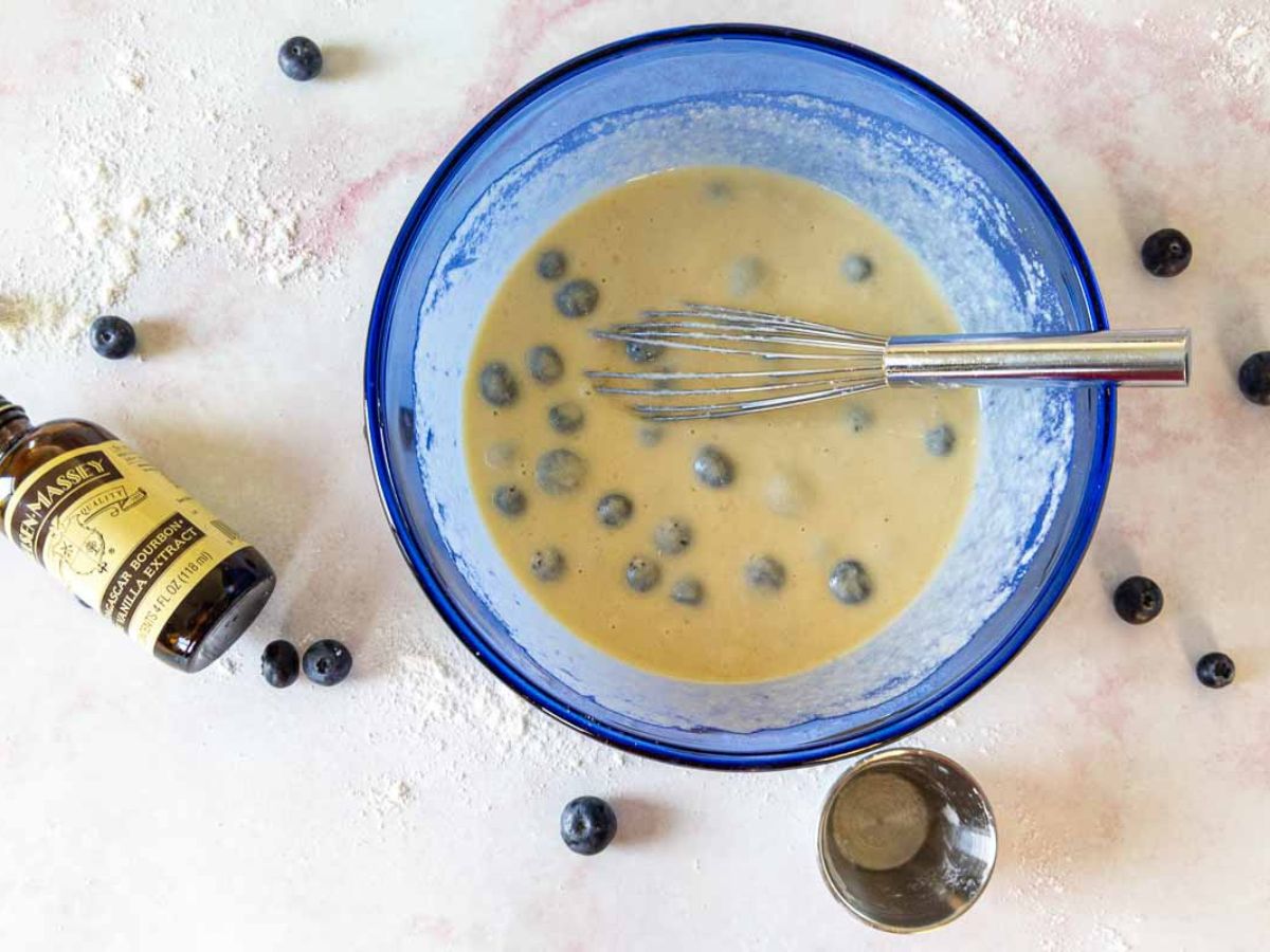 A blue mixing bowl filled with blueberry pancake batter and a metal whisk sits on a floured surface. Nearby are blueberries, a small metal cup, and a bottle of vanilla extract lying on its side.