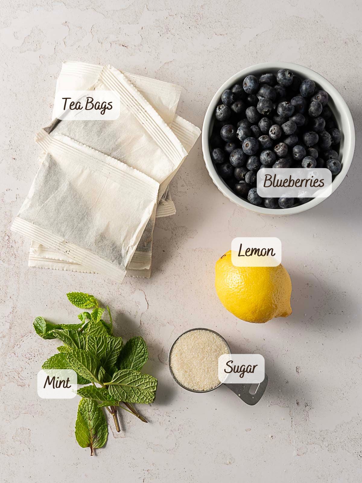 A flat lay of ingredients labeled as tea bags, a bowl of blueberries, a lemon, sugar in a measuring cup, and a bunch of mint on a light countertop.
