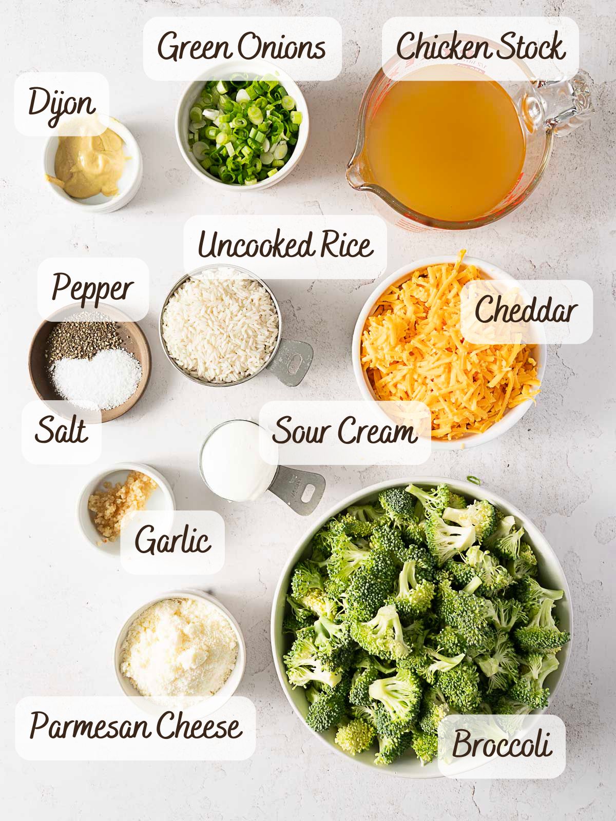 Bowls and containers of labeled ingredients for a Cheesy Broccoli Rice Casserole, including broccoli, cheddar, rice, sour cream, chicken stock, green onions, Parmesan, garlic, Dijon mustard, salt, and pepper.