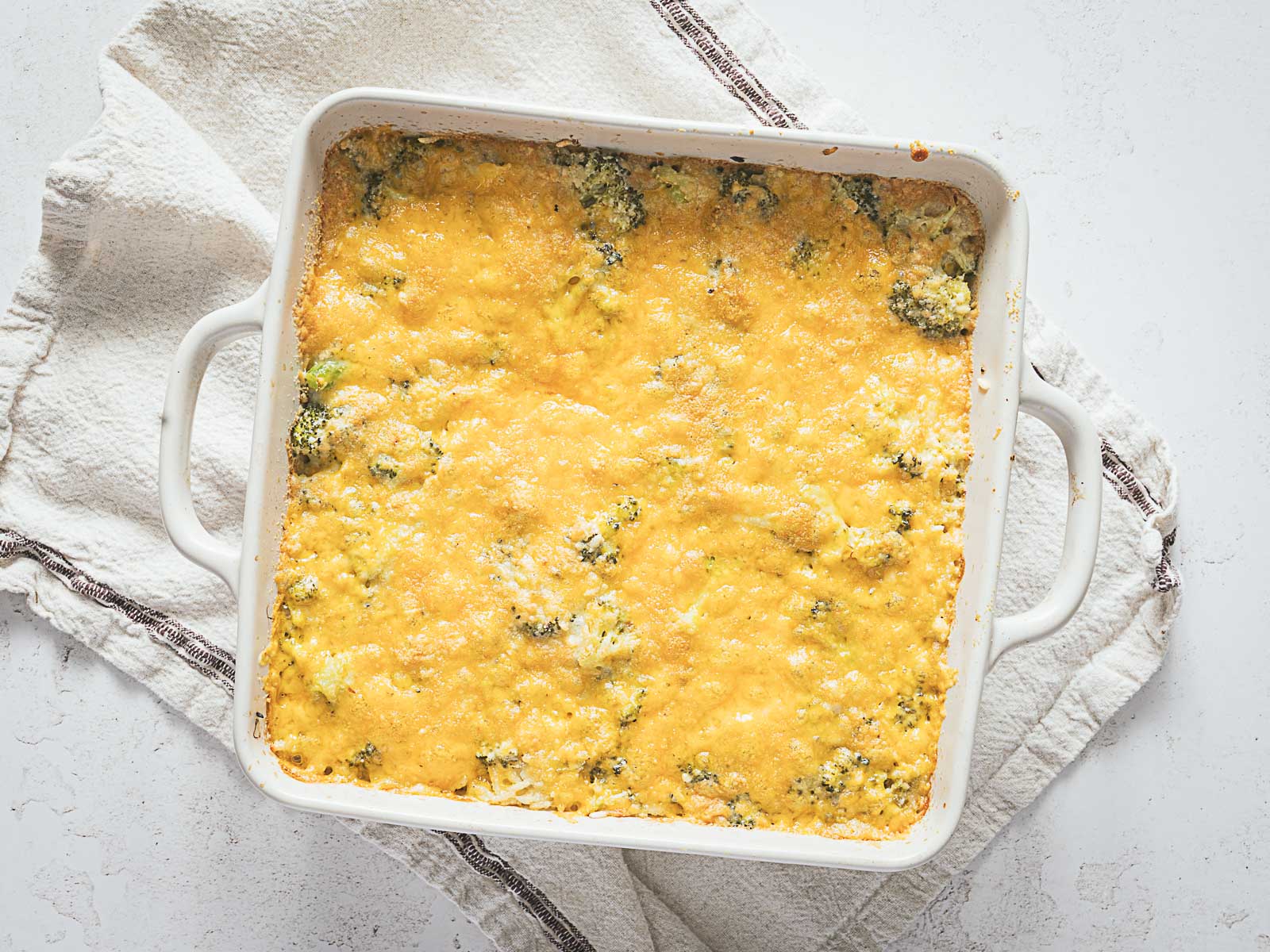 A square white baking dish filled with Cheesy Broccoli Rice Casserole, its golden melted cheese topping inviting and warm, rests on a folded kitchen towel.