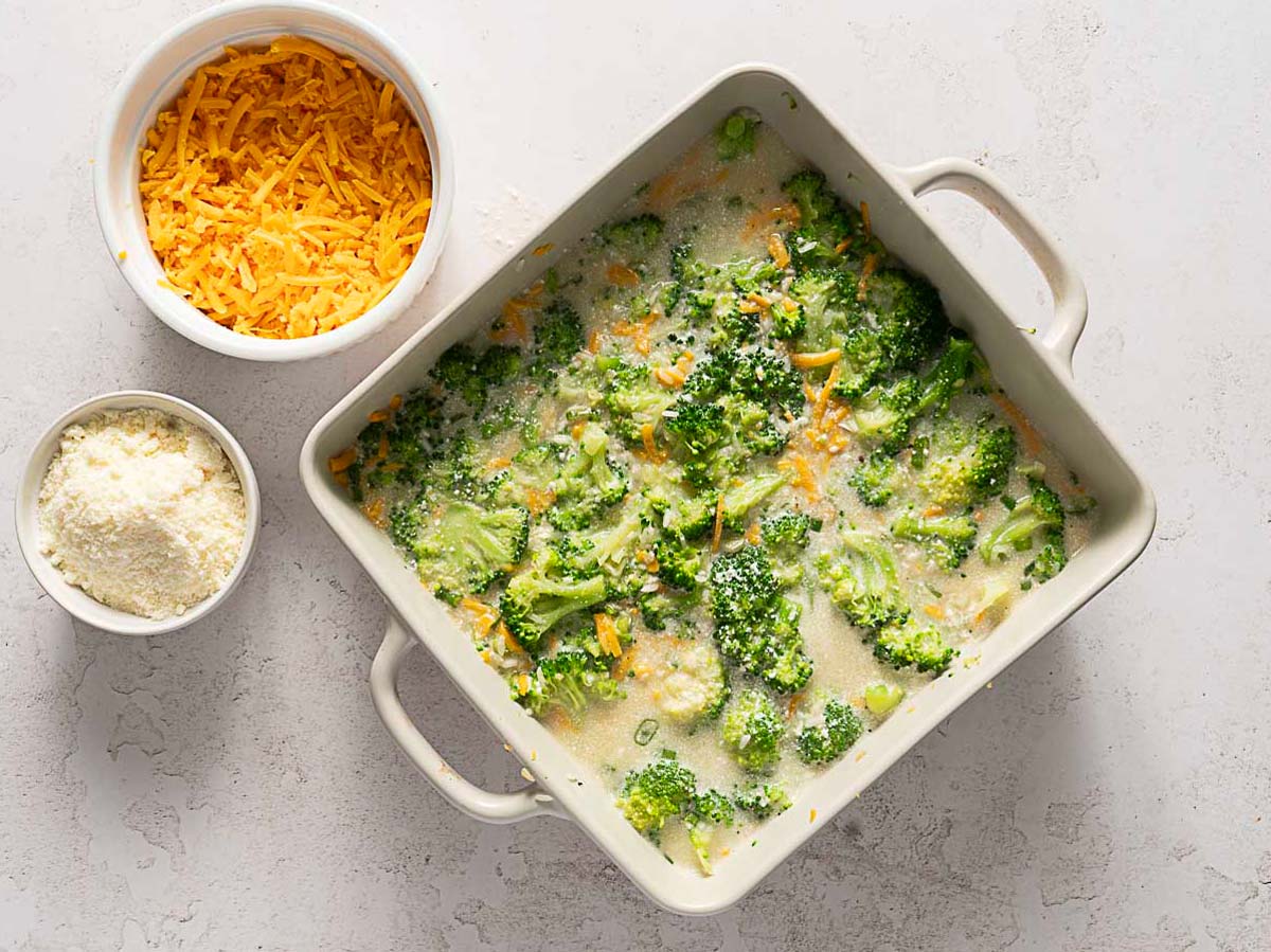 A Cheesy Broccoli Rice Casserole filled with broccoli in a creamy sauce sits on a counter next to bowls of shredded cheddar and grated parmesan cheese.